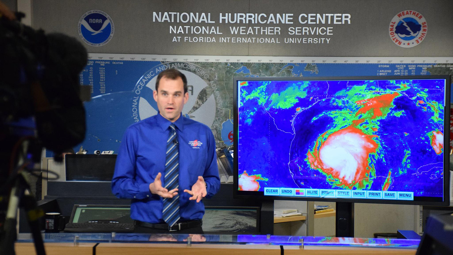 Michael Brennan stands next to a satellite image of a hurricane on a screen in the National Hurricane Center