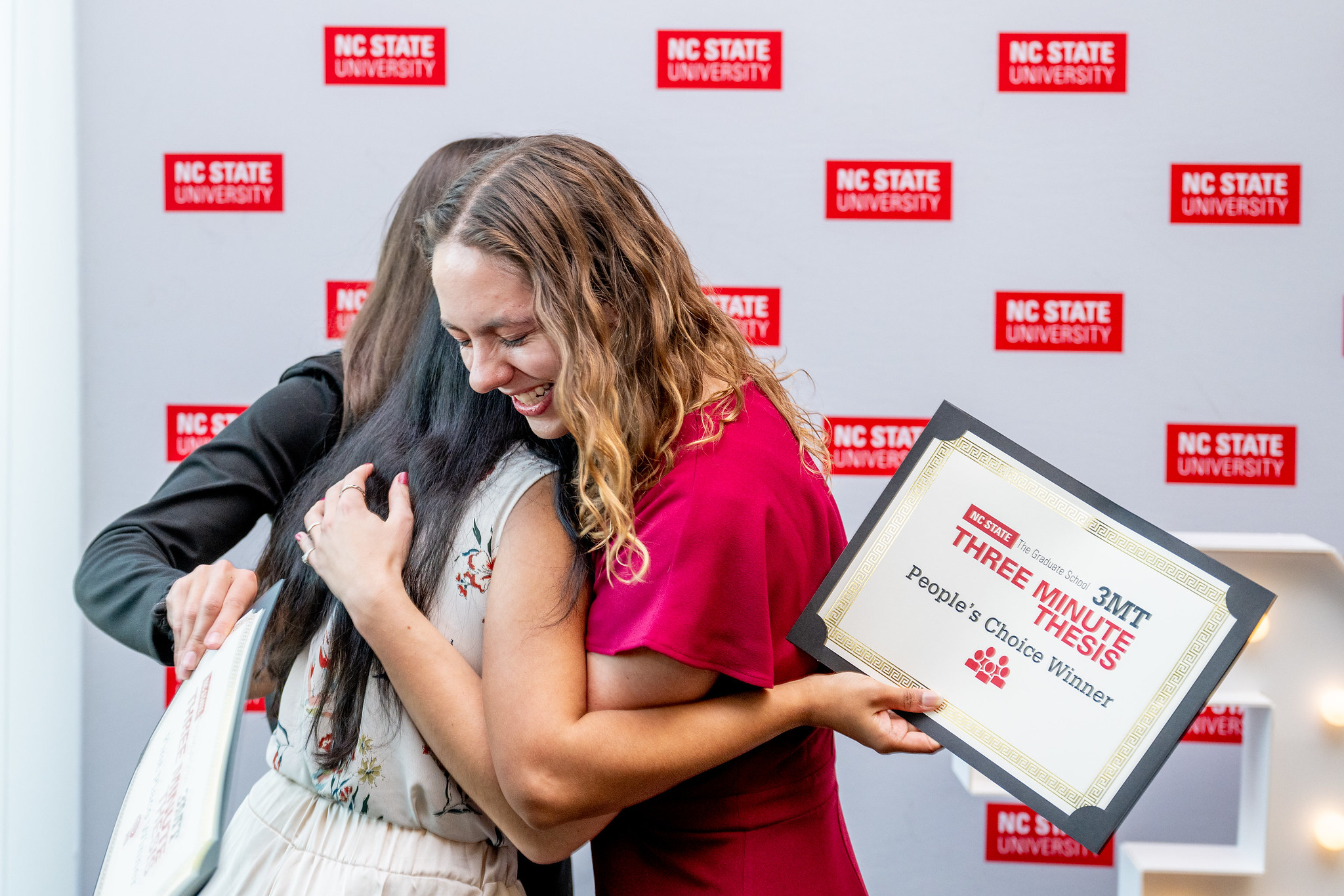 Alyssa Griffin, wearing red, smile and hugs two other winners of the Three Minute Thesis challenge