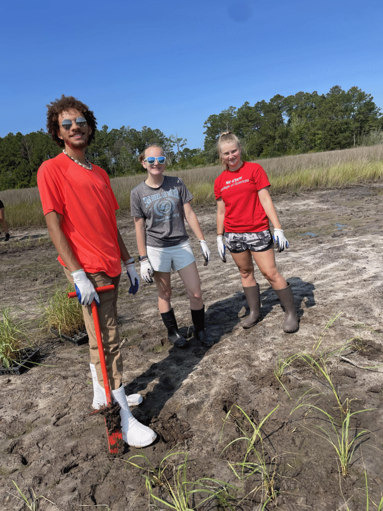 Three students smiling while holding an orange dibble bar in the field