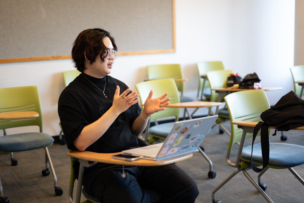 A student is speaking and gesturing with hands while seated  at a desk with a laptop